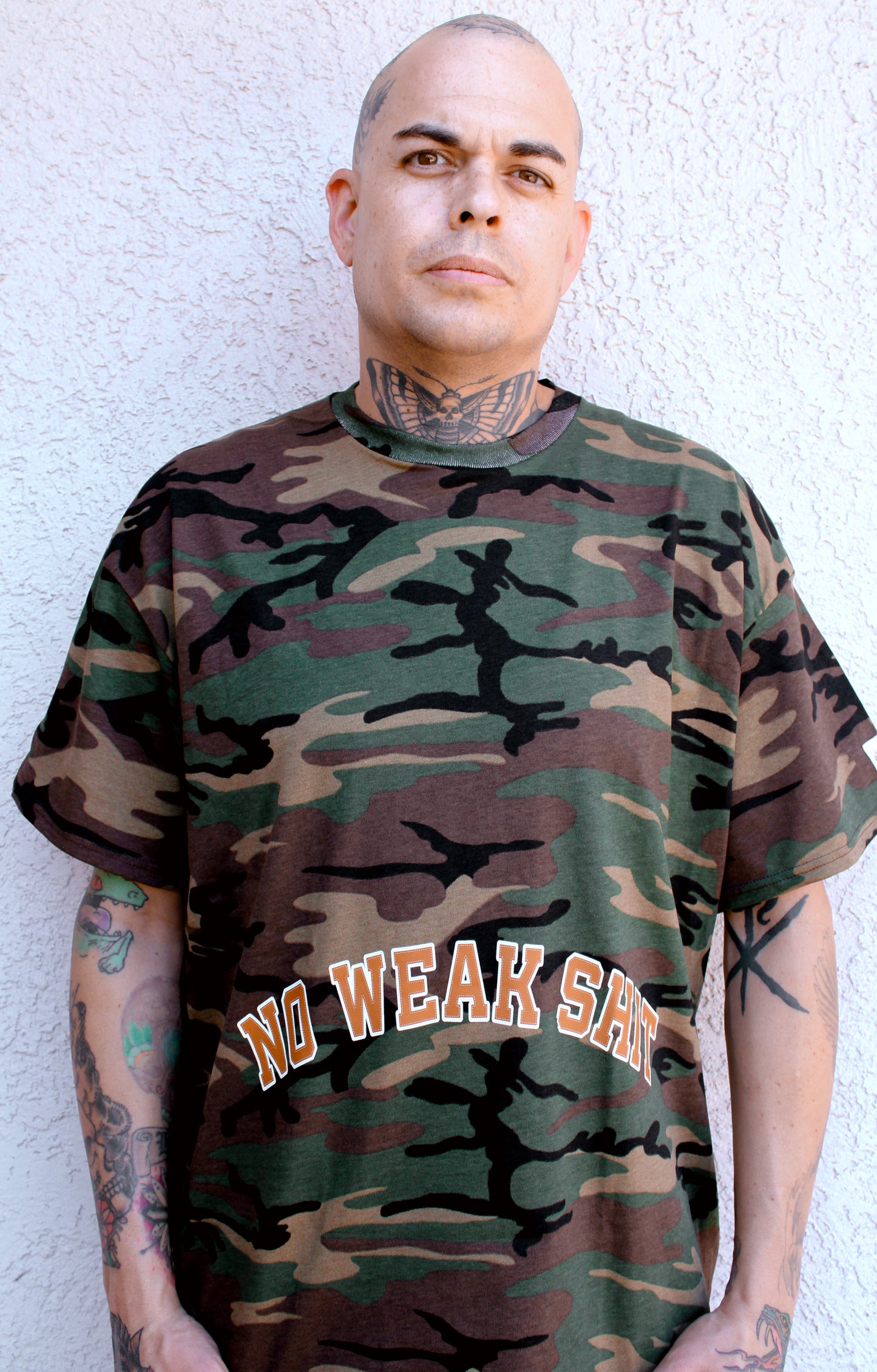 Person wearing a camouflage t-shirt with 'NO WEAK SHIT' text against a white wall.