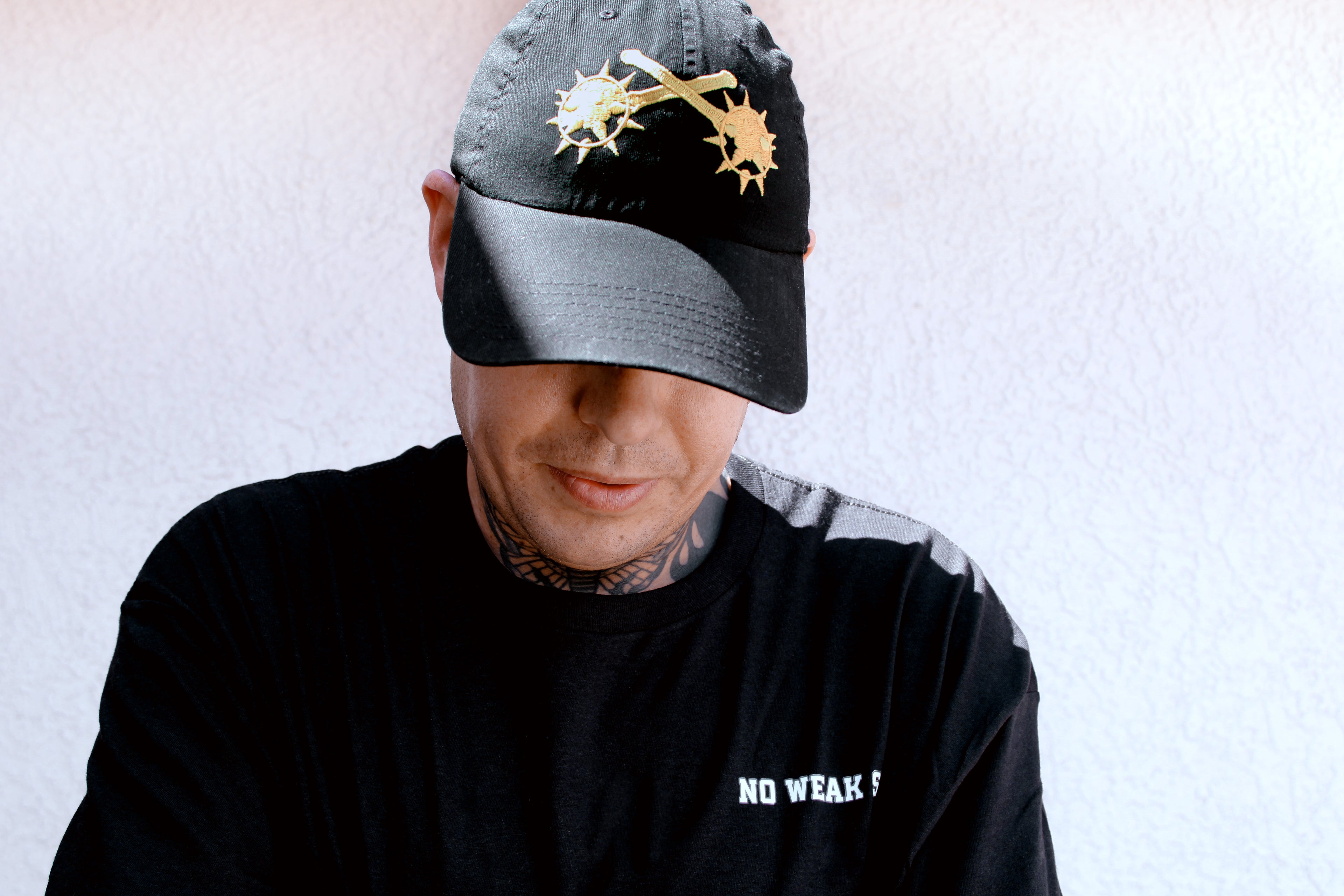 Person wearing a black cap with a gold embroidered design of crossed mace brand logo on a light gray background