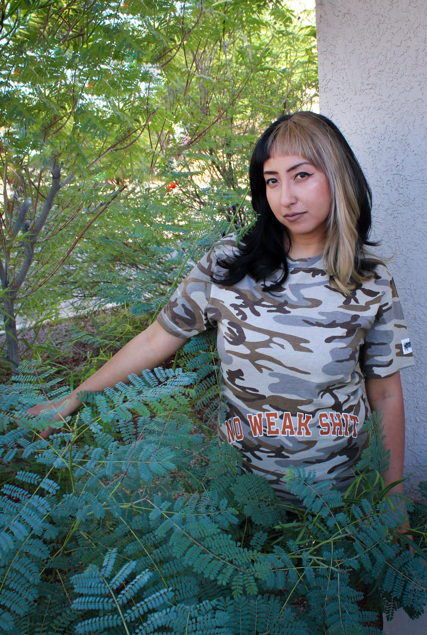 Woman wearing a camouflage t-shirt with text, standing outdoors near greenery.