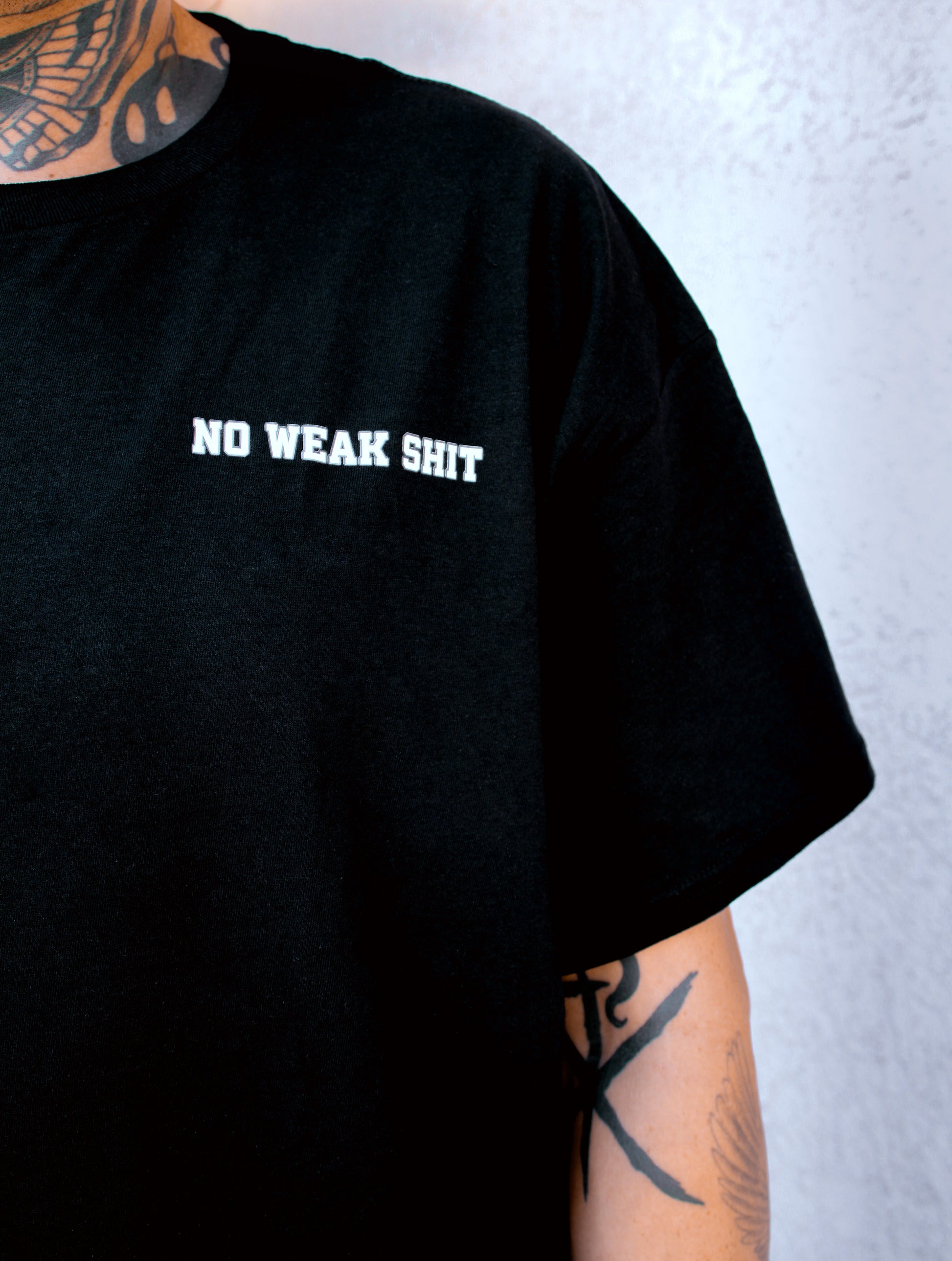Black t-shirt with 'NO WEAK SHIT' text worn by a person with tattoos on a light gray background